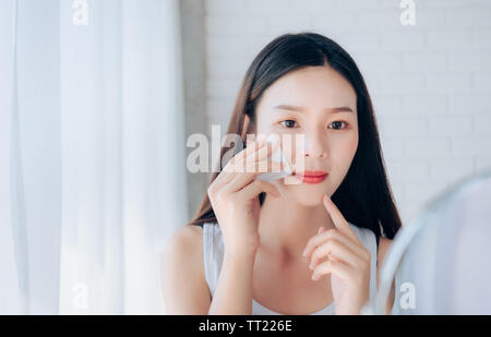 Junge Schönheit asiatische Frau Reinigung Gesicht mit Baumwolle Ihr auf Spiegel klares Gesicht Hautpflege und Lächeln Morgen in Weiß Schlafzimmer. Stockfoto