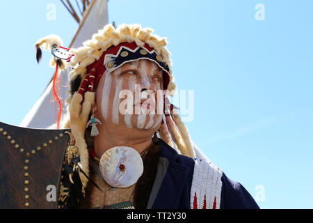 Ein Mann in den Krieg malen und Kleidung eines Nordamerikanischen Indianer steht mit der Waffe in der Nähe von einem Wigwam gegen den blauen Himmel. Wiederaufbau des native american Stockfoto