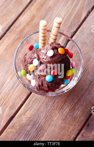 Schokoladeneis mit bunten Bonbons und Waffelröllchen in Glas Schüssel, auf Holz- Hintergrund Stockfoto