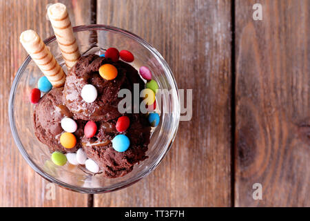 Schokoladeneis mit bunten Bonbons und Waffelröllchen in Glas Schüssel, auf Holz- Hintergrund Stockfoto