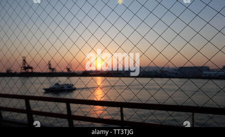 Zaun, mit unscharfen Hintergrund von Meer mit Schiff, skizziert von port Cargo Cranes und Gebäude, gegen die untergehende Sonne mit Twilight in den Himmel, Stockfoto