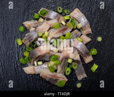 Schichten von gesalzenem Fleisch ohne Knochen, die in der Wildnis gefangen Atlantischer Hering garniert mit gehackten Frühlingszwiebeln Frühlingszwiebeln. Schwarz Naturstein Hintergrund. Clupea harengus festgelegt. Stockfoto