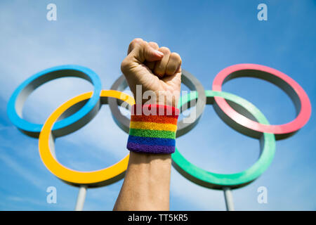 LONDON - Mai 4, 2019: eine Hand tragen Gay Pride Regenbogen farbigen Armband macht einen feierlichen Faust vor der olympischen Ringe unter blauem Himmel stehen. Stockfoto