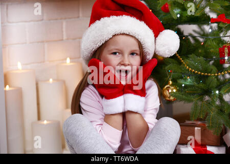 Kleines Mädchen in Santa Hut und Handschuhe sitzen in der Nähe von Tanne am Kamin mit Kerzen Hintergrund Stockfoto