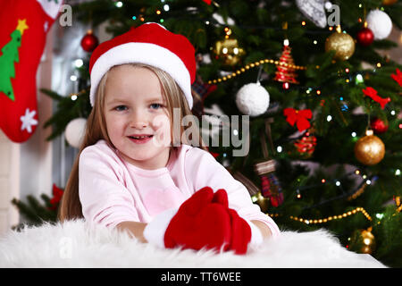 Kleines Mädchen in Santa Hut und Handschuhe liegen auf Fell Teppich auf Weihnachtsbaum Hintergrund Stockfoto