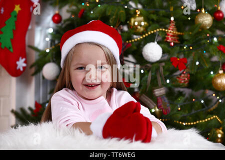 Kleines Mädchen in Santa Hut und Handschuhe liegen auf Fell Teppich auf Weihnachtsbaum Hintergrund Stockfoto