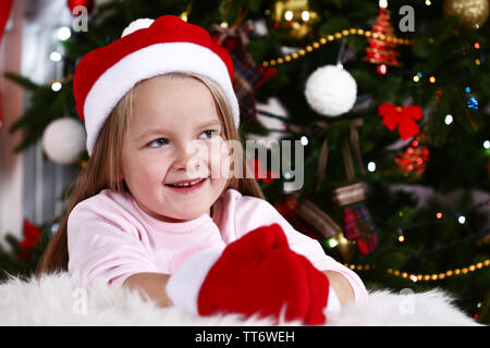 Kleines Mädchen in Santa Hut und Handschuhe liegen auf Fell Teppich auf Weihnachtsbaum Hintergrund Stockfoto