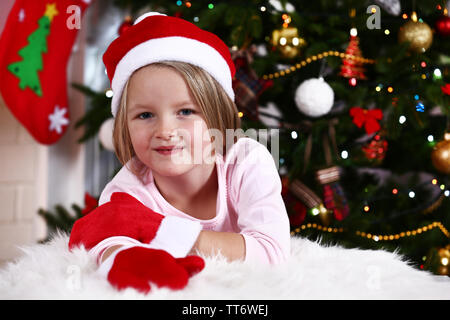 Kleines Mädchen in Santa Hut und Handschuhe liegen auf Fell Teppich auf Weihnachtsbaum Hintergrund Stockfoto