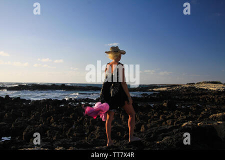 Isolierte blonde Frau mit Strohhut, Batik Kleid, winken Schal im Wind stehend auf Felsen am Meer in der Abendsonne - Fuerteventura, El Cotillo Stockfoto
