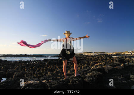 Isolierte blonde Frau mit Strohhut, Batik Kleid, winken Schal im Wind stehend auf Felsen am Meer in der Abendsonne - Fuerteventura, El Cotillo Stockfoto