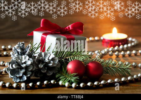 Weihnachten Zusammensetzung: ein Geschenk mit roter Schleife silber Kegel, rote Kugeln und ein Fir Tree Branch - brennende Kerze im Hintergrund und Schneeflocken umhüllt Stockfoto