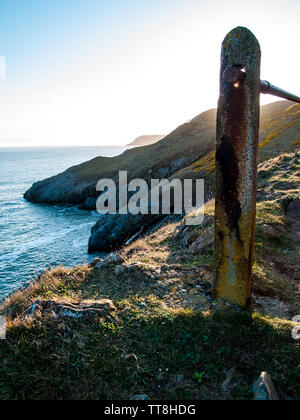 Eine untergehende Sonne scheint durch ein Loch auf einem Geländer auf dem Küstenweg zwischen Langland Bay und Caswell Bay, Gower, Wales, Großbritannien. Stockfoto