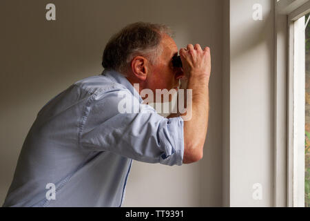 Ältere zerzaust aussehenden Mann starrte aus dem Fenster durch ein Fernglas Stockfoto