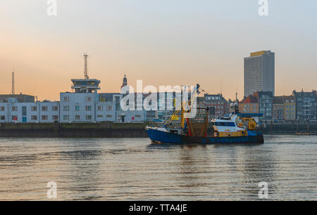 Fischerboot in den Hafen von Oostende (Ostende) Stadt in der Abenddämmerung mit der Skyline und dem höchsten Wolkenkratzer im Hintergrund, Belgien. Stockfoto