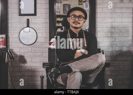 Portrait von zuversichtlich Stylist mann Friseur im Vintage barbershop Stuhl mit gekreuzten Arm vor und auf der Kamera sitzen Stockfoto