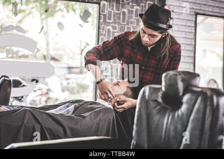 Nahaufnahme Portrait von asiatischen Friseur mit langen Haaren Bart rasieren, seine Kunden bei der Barbershop Stuhl, während seine männlichen Kunden bei Friseur Stuhl entspannt Stockfoto