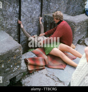 1960er Jahre, historisch, eine junge Frau in einem Minirock, die auf einem Teppich liegt und den blarney-Stein, den berühmten alten Kalkstein in Blarney Castle, Cork, Irland, küsst. Sie wird von einem Helfer, einem jungen Mann, festgehalten, da es aufgrund seiner Lage unter den Zinnen nur möglich ist, kopfüber zu liegen. Der Stein wurde in einen Turm der Burg eingesetzt, als er 1446 aus einem Donjon umgebaut wurde, und der Legende nach gibt „Küssen des blarney-Steins“ die „Gabe des Gaben“ oder „große Beredsamkeit“. Es ist eine der wichtigsten Touristenattraktionen in Südirien. Stockfoto