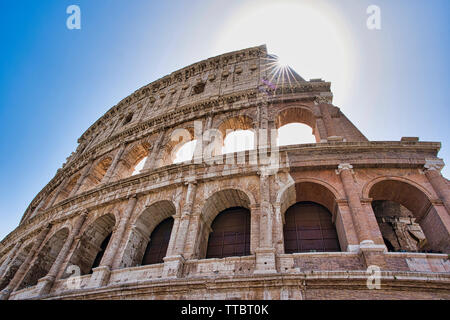 Flacher Blick auf das Kolosseum in Rom, Italien, mit Sonnenschein über alten Bögen vor einem klaren blauen Himmel. Stockfoto
