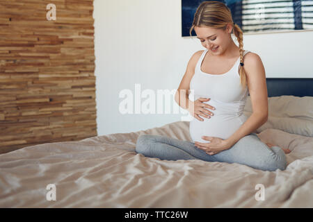 Glückliche junge schwangere Frau Verklebung mit ihrem ungeborenen Kind sitzen auf einem Bett mit Ihrem Babybauch mit ihren Händen nach unten schauen. Stockfoto