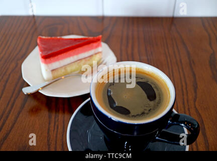 Tasse heißen Kaffee auf Holztisch mit einer Scheibe Himbeermousse Kuchen im Hintergrund Stockfoto