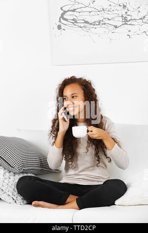 Junge Frau sitzt auf dem bequemen Sofa mit Schale auf Kaffee in der Hand und sprechen über Mobiltelefon Stockfoto