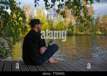 Reifer Mann angeln vom hölzernen Pier in der Nähe von Ferienhaus am See in Finnland im Sommer Stockfoto