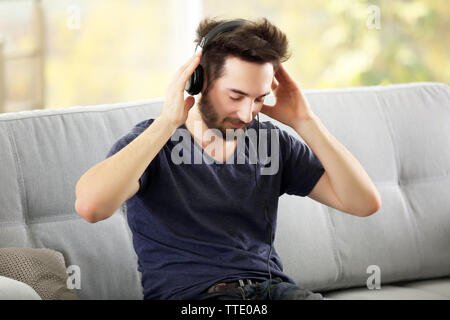 Schöner Mann hört emotional Musik mit Kopfhörern auf grau Sofa im Zimmer Stockfoto