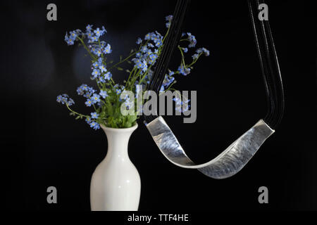 Gebogen, glänzendes Metall hängen an Schwarze dünne Schnürsenkel und einen Strauß Vergissmeinnicht in eine weiße Vase. Stockfoto