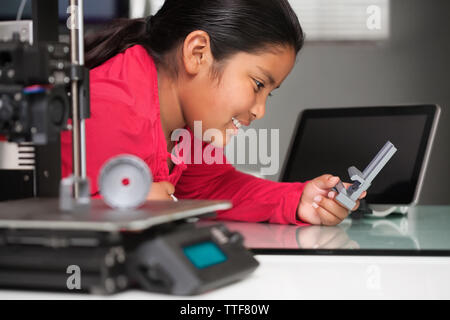 Eine lächelnde junge Mädchen ist glücklich, Ihre 3D-gedruckten Modell zu sehen und hält es in der Hand, mit einem 3D-Drucker im Vordergrund und im Hintergrund. Stockfoto