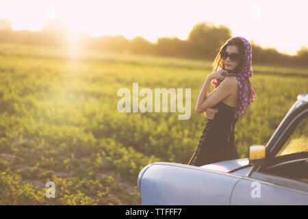 Portrait von Frau mit Sonnenbrille steht mit dem Auto auf der Wiese gegen Himmel bei Sonnenuntergang Stockfoto