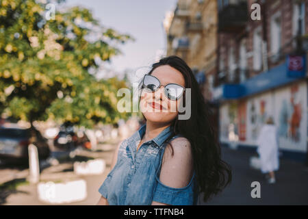 Lächelnden jungen Frau Sonnenbrille tragen, während in der Stadt stehend Stockfoto