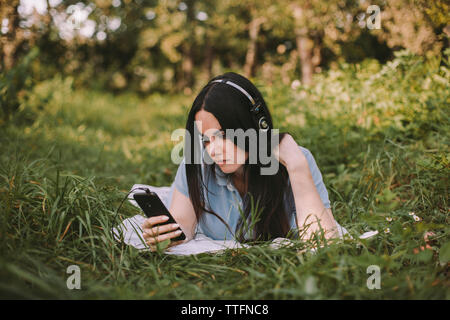 Frau mit langem Haar Musik hören, während Sie Mobiltelefon auf Wiese im Garten Stockfoto