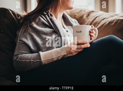 Zugeschnittenes Bild der Frau mit Becher mit Wort auf es auf der Couch entspannen. Stockfoto