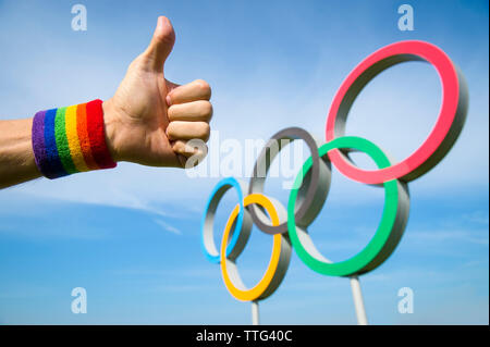 LONDON - Mai 4, 2019: eine Hand tragen Gay Pride Regenbogen farbige Bändchen gibt einen "Daumen-nach-oben-Geste vor der Olympischen Ringe in den blauen Himmel. Stockfoto