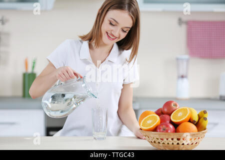 Junge Frau gießt Wasser aus der Kanne in Glas in der Küche Stockfoto