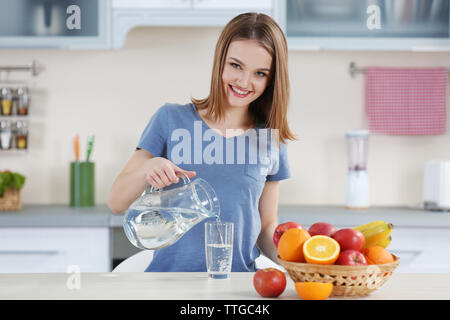 Junge Frau gießt Wasser aus der Kanne in Glas in der Küche Stockfoto