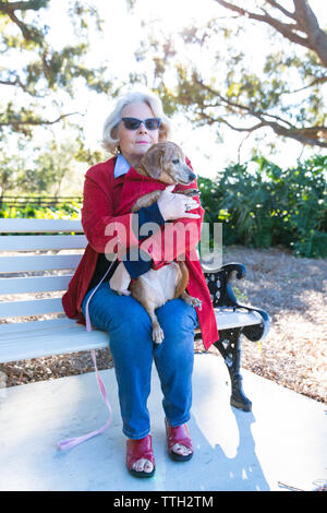 Eine ältere Frau sitzt im Freien auf einer Bank in einem Park mit ihrem Hund. Stockfoto