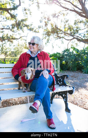 Eine ältere Frau sitzt im Freien auf einer Bank in einem Park mit ihrem Hund. Stockfoto