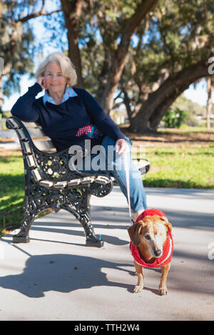 Eine ältere Frau sitzt im Freien auf einer Bank in einem Park mit ihrem Hund. Stockfoto