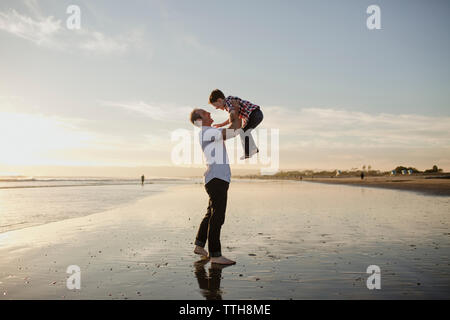 Verspielt Vater anheben Sohn beim Spielen am Strand gegen Himmel bei Sonnenuntergang Stockfoto