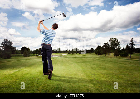 Volle Länge Rückansicht des Menschen schwingen Golf Club auf Feld gegen bewölkter Himmel Stockfoto