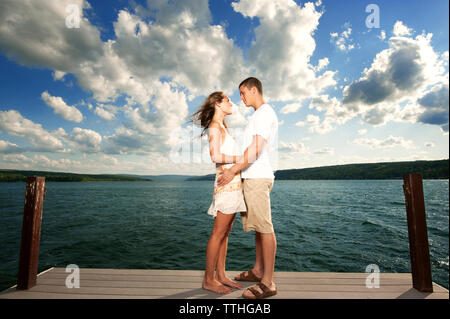 Seitenansicht des jungen Paares sich einander beim Stehen auf Steg über den See gegen Sky Stockfoto