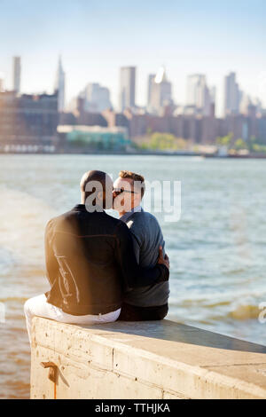 Rückansicht der homosexuellen Männer küssen, beim Sitzen auf der Stützmauer am Fluss Stockfoto