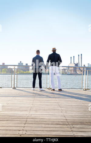 Rückansicht der homosexuellen Männer halten sich an den Händen und stehen auf Pier gegen den klaren Himmel Stockfoto