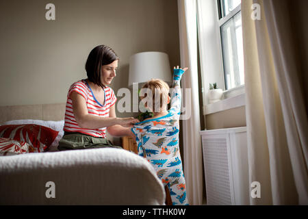 Mutter dressing Sohn beim Sitzen auf dem Bett im Zimmer Stockfoto