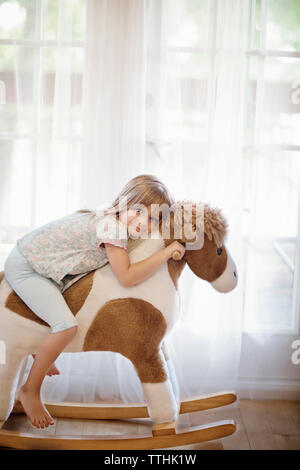 Portrait von lächelnden Mädchen auf Schaukelpferd gegen die Vorhänge zu Hause sitzen Stockfoto