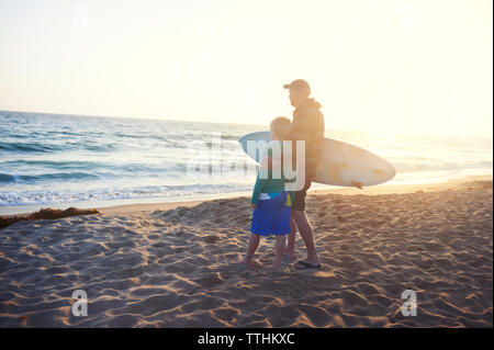 Älterer Mann, Surfbrett, während mit Enkel am Strand stehen Stockfoto