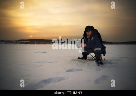 Mann Eisfischen auf dem zugefrorenen See gegen Himmel bei Sonnenuntergang Stockfoto