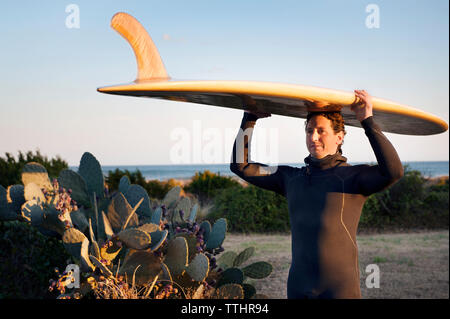 Surfer, Surfbrett auf dem Kopf stehend auf der Wiese gegen den klaren Himmel Stockfoto