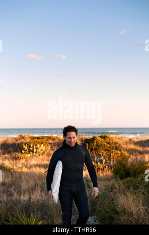 Surfer Surfbrett Durchführung beim Gehen auf Wiese gegen Sky Stockfoto